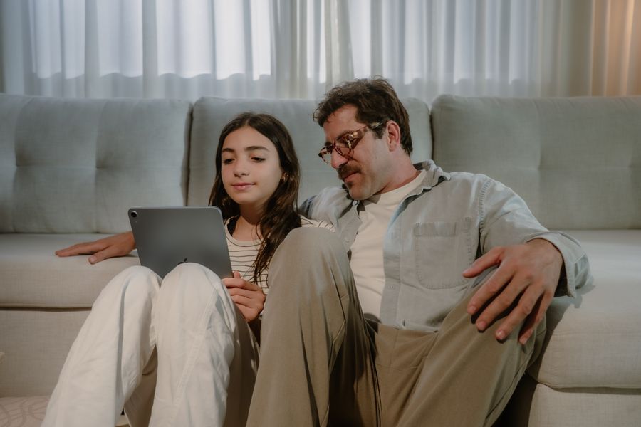 Pai e filha sentados no chão da sala, encostados no sofá, lendo uma história no tablet.