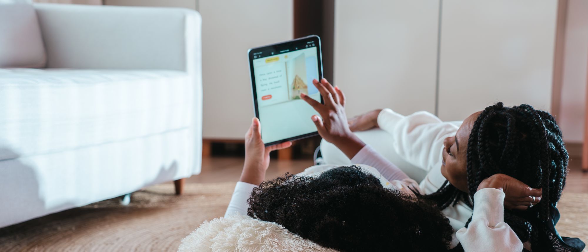 Mãe e filha deitadas nas almofadas da sala, lendo juntas uma história no tablet.