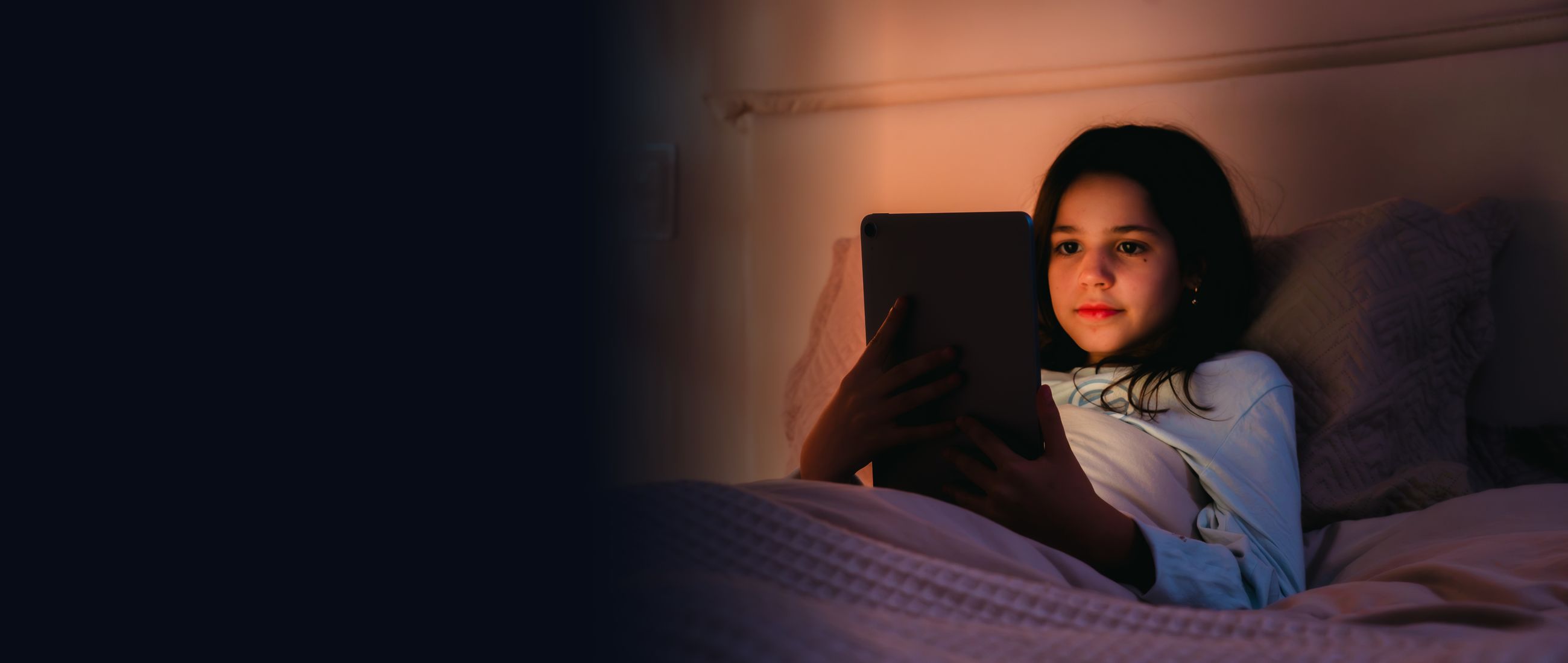 Menina deitada na cama à noite, com o rosto iluminado pelo tablet enquanto lê uma história no Imagine.