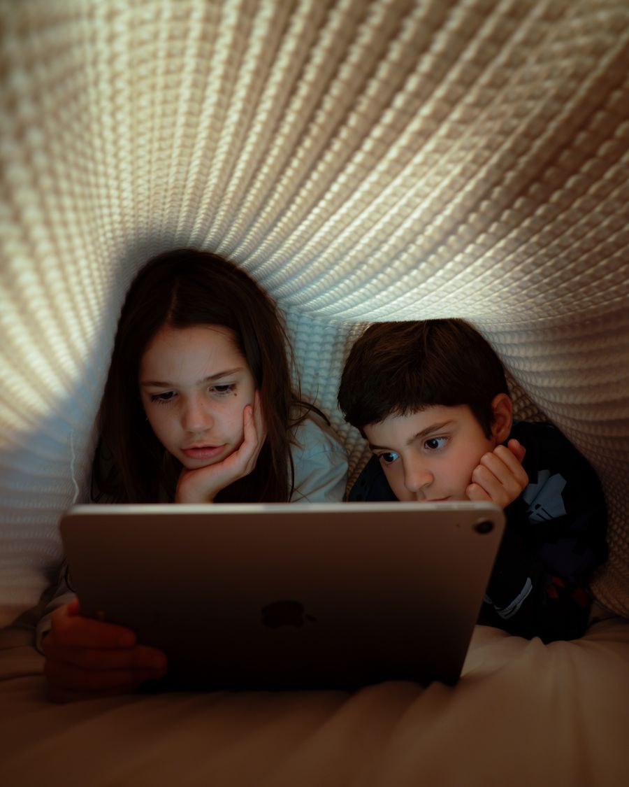 Dois irmãos dentro de uma cabana de cobertor, lendo uma história no tablet antes de dormir.